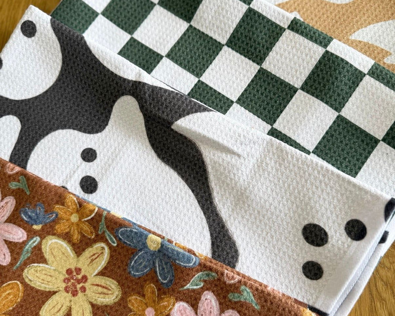 Tidy Towel October kitchen towels: ghost, green check and autumn floral waffle towels stacked on a wood table