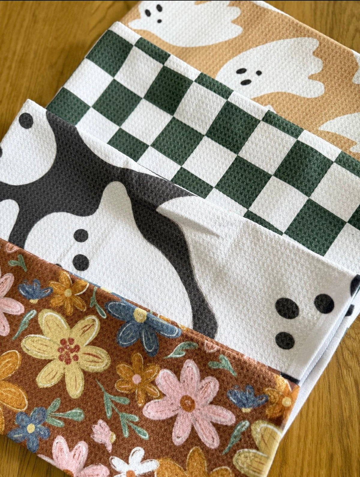 Tidy Towel October kitchen towels: ghost, green check and autumn floral waffle towels stacked on a wood table