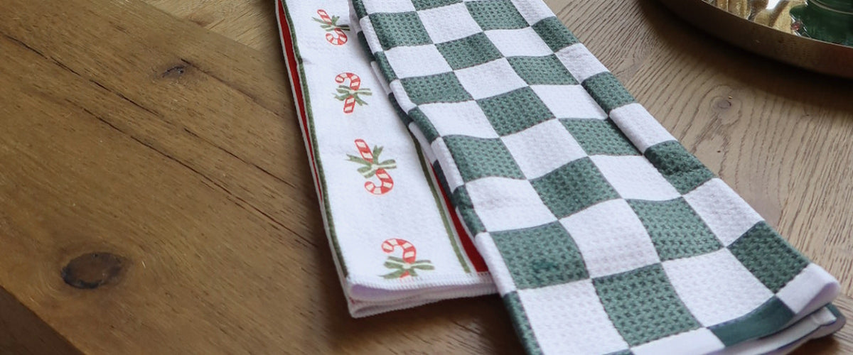 Christmas kitchen towels (Candy Cane stripe and green check) folded on a wooden table by a Christmas tree